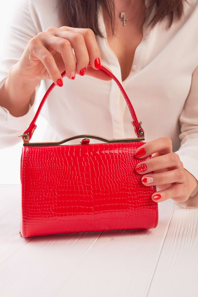 A person with red painted nails holds a small, textured red handbag on a white wooden surface—an elegant accessory to prevent any unexpected wardrobe malfunctions.